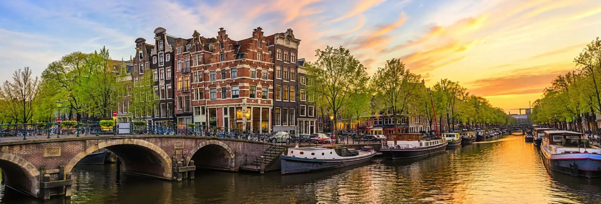 Sunset view of Amsterdam's Canal Ring with historic canal houses, arched bridge, and boats along the water.
