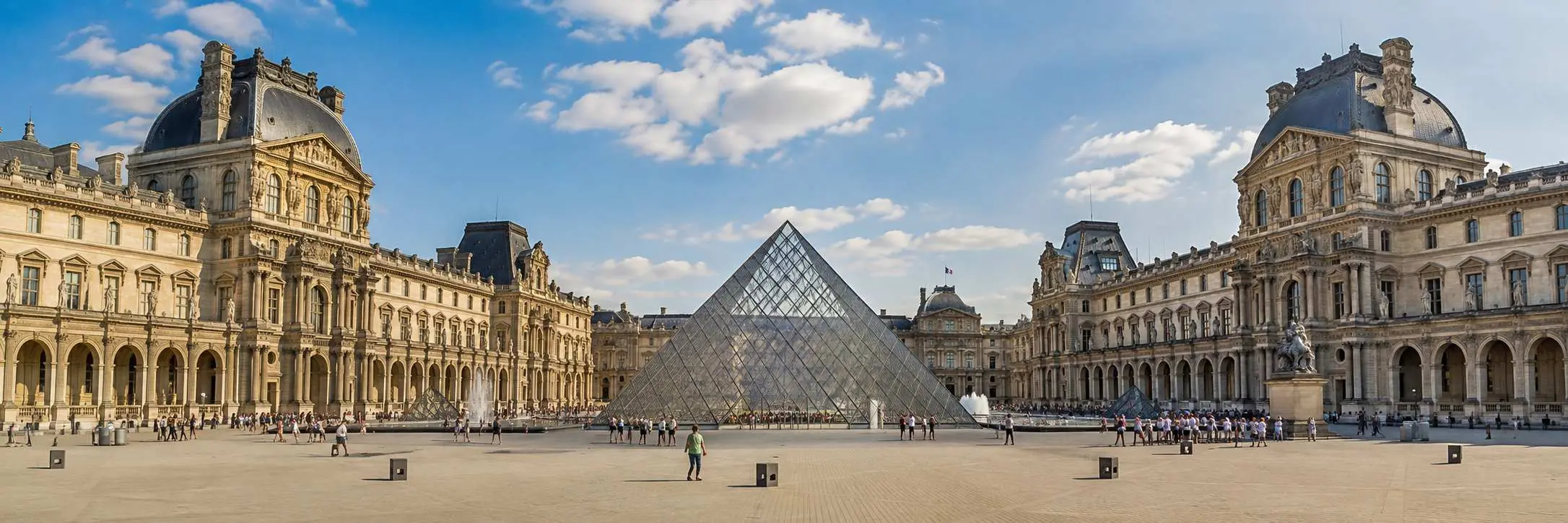 Louvre Museum in Paris with its iconic glass pyramid and historic palace buildings under a bright blue sky in a panorami