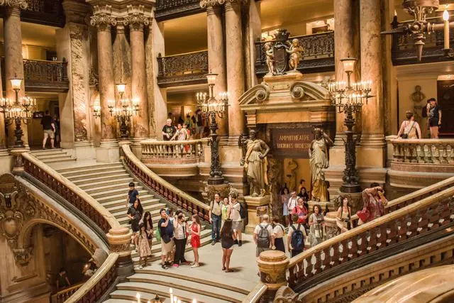 The Grand Staircase inside Palais Garnier with ornate marble columns, chandeliers, and visitors.