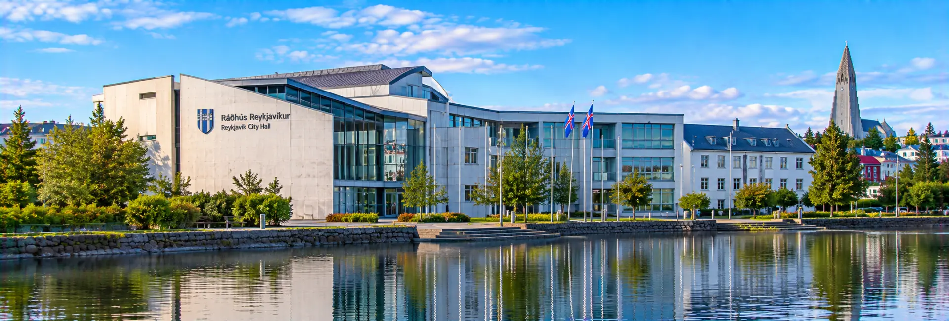 Reykjavík City Hall beside a reflecting pond with Icelandic flags and Hallgrímskirkja