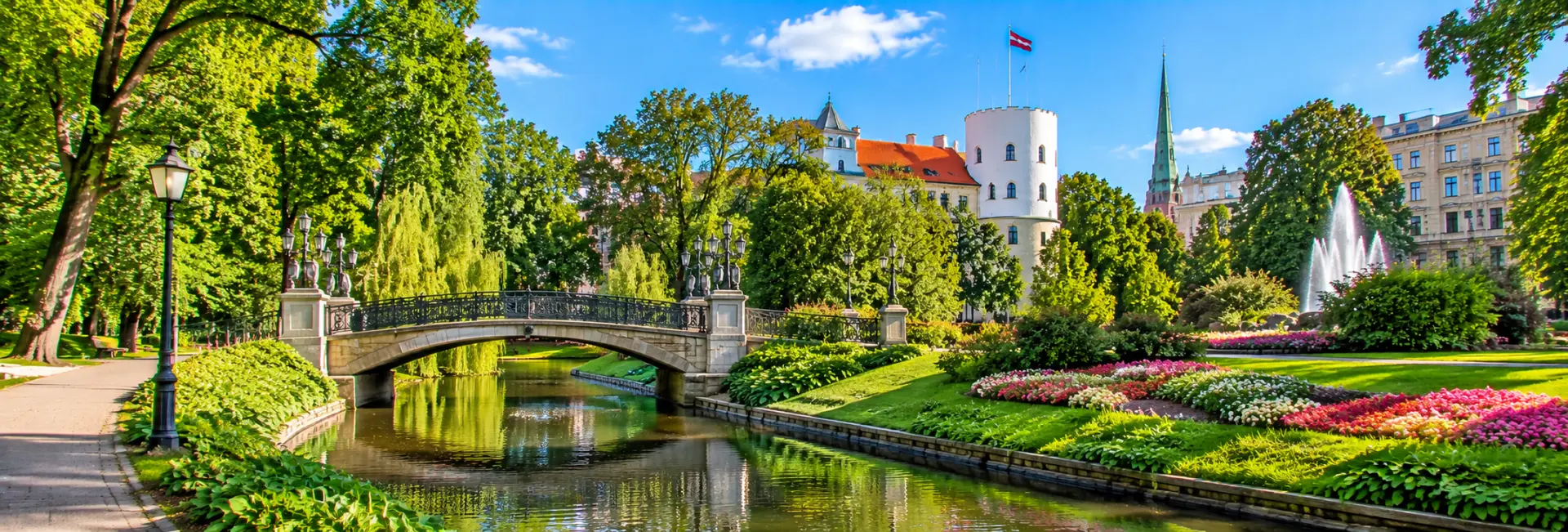 Bastejkalns Park in Riga, Latvia, featuring a scenic canal, pedestrian bridge, colorful flowerbeds and lush greenery.