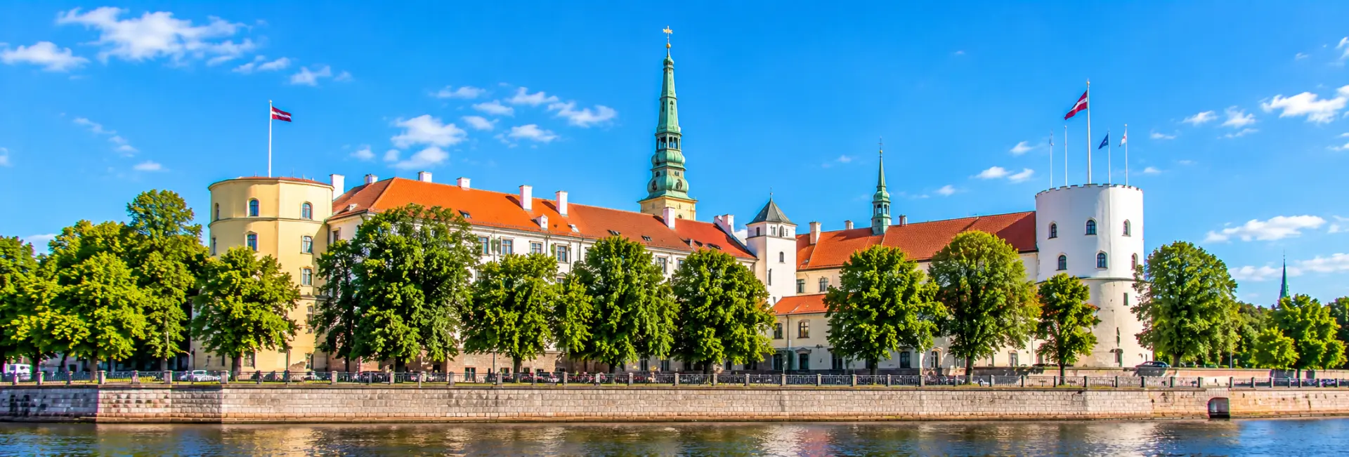 Riga Castle in Latvia, featuring historic towers, red-tiled roofs and Latvian flags along the Daugava River.
