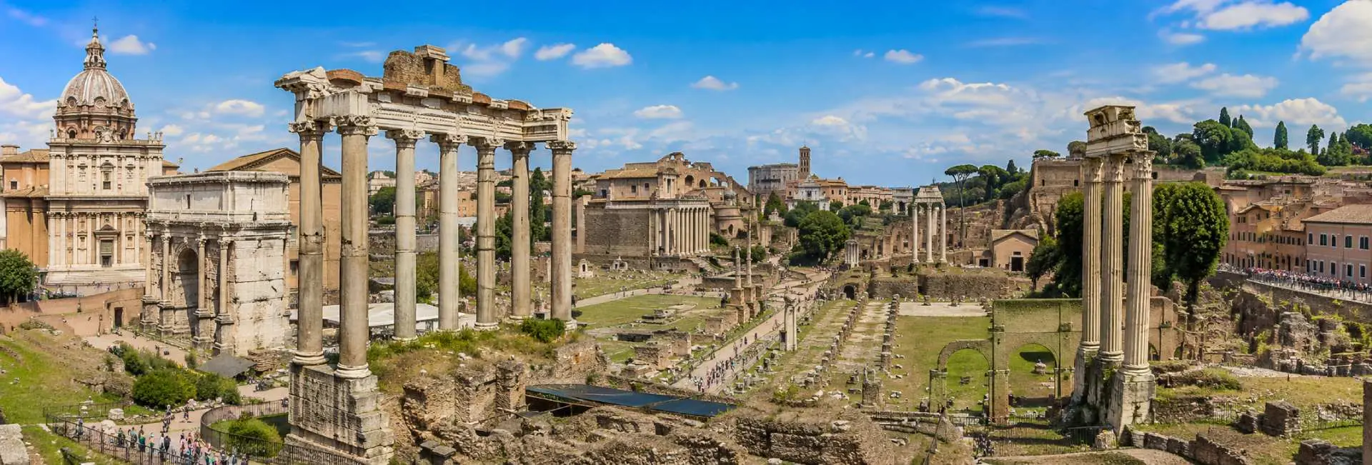 The Roman Forum ruins in Rome with ancient columns, temples, and historic buildings under a blue sky.