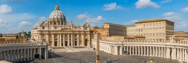 Panoramic daytime view of Vatican City featuring St. Peter's Basilica and St. Peter's Square under a blue sky.
