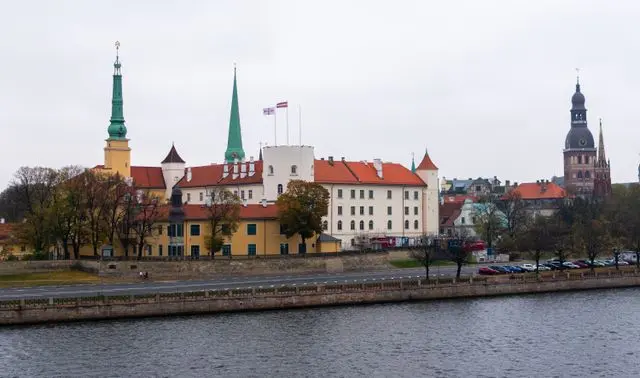 Panoramic view of Riga Castle, historic church towers, and colorful buildings along the Daugava River in Latvia.