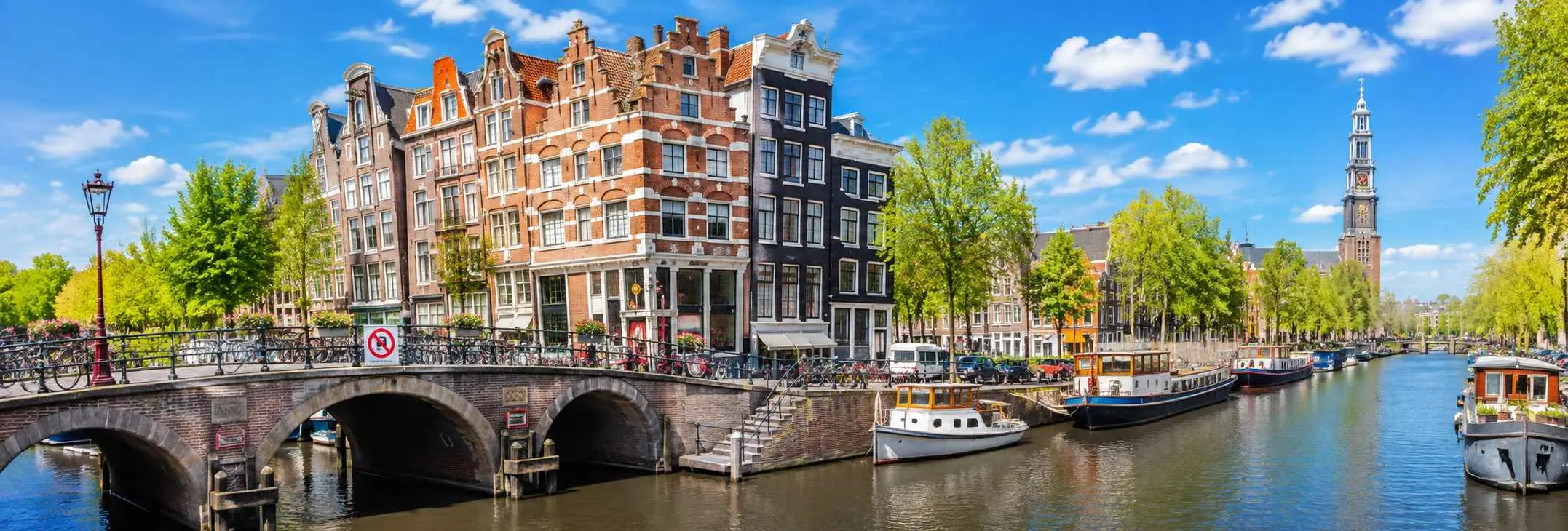 Panoramic view of Amsterdam featuring historic canal houses, bridges, boats, and scenic canals under a clear blue sky.