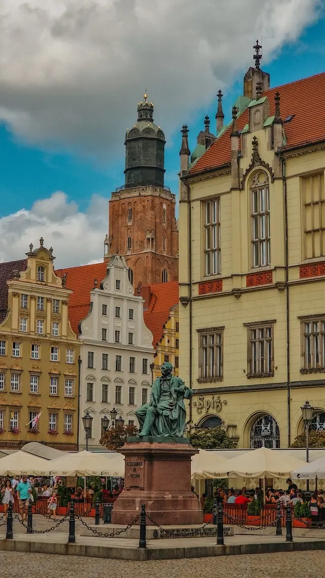 Historic European city square with colorful buildings, a brick church tower, and a seated statue