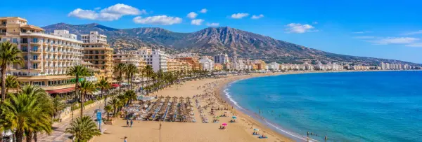 Torremolinos, Spain, with a sunny Mediterranean beach, palm-lined promenade, resort buildings, and mountain backdrop